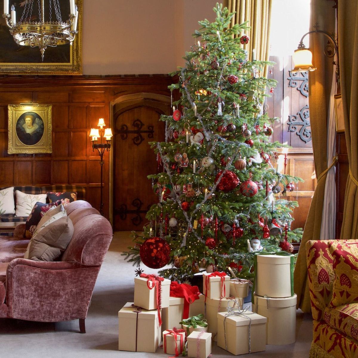 A decorated Christmas tree in a warm, wood-paneled living room with wrapped presents underneath. The tree is adorned with red and silver ornaments, and the room features a pink velvet armchair, a patterned armchair, and warm lighting.