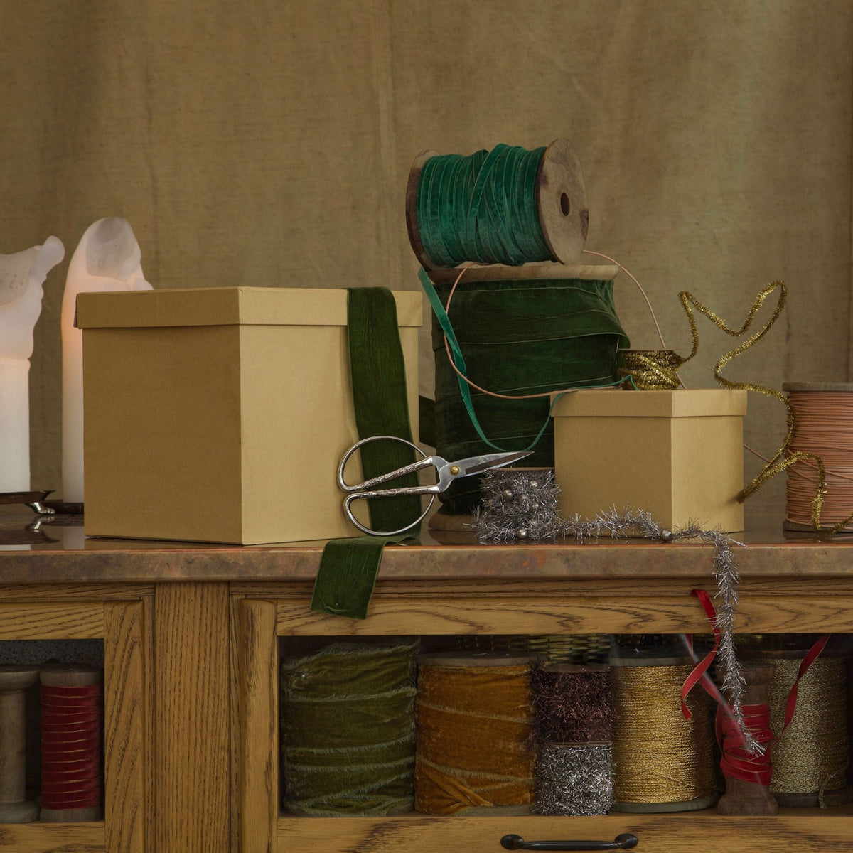 A styled arrangement of gift boxes, spools of ribbon, and silver scissors on a wooden counter against a textured beige backdrop.
