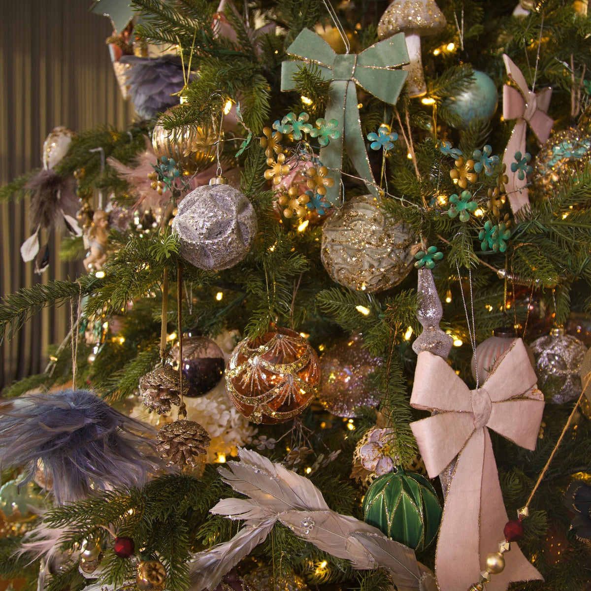 A decorated Christmas tree with pastel pink and sage green ornaments, including large fabric bows, glittery baubles, feathers, and warm string lights.
