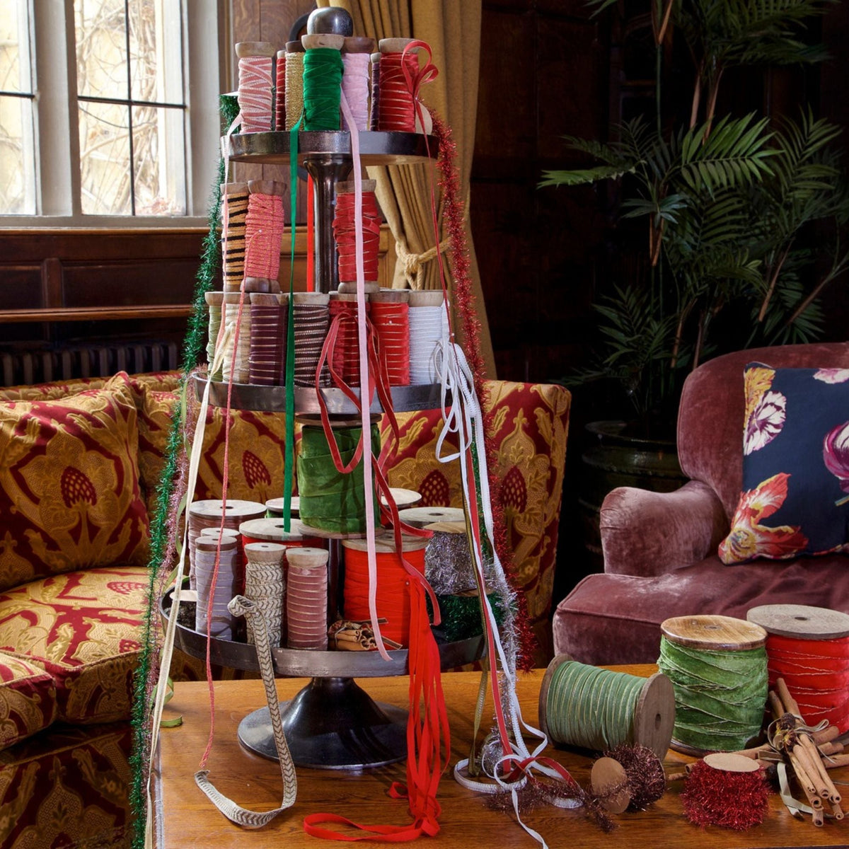 A multi-tiered black metal spindle rack filled with colorful spools of thread, ribbon, and cord, styled on a wooden table in a cozy, vintage-inspired room with patterned armchairs and a plant in the background.