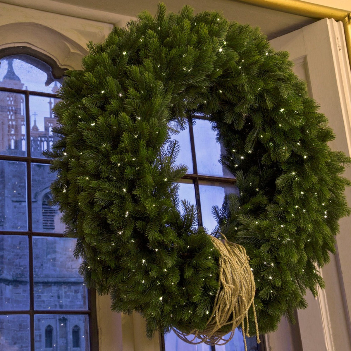 A large green pine wreath with small white string lights and a natural jute rope bow, hanging on a window with a stone building visible outside.