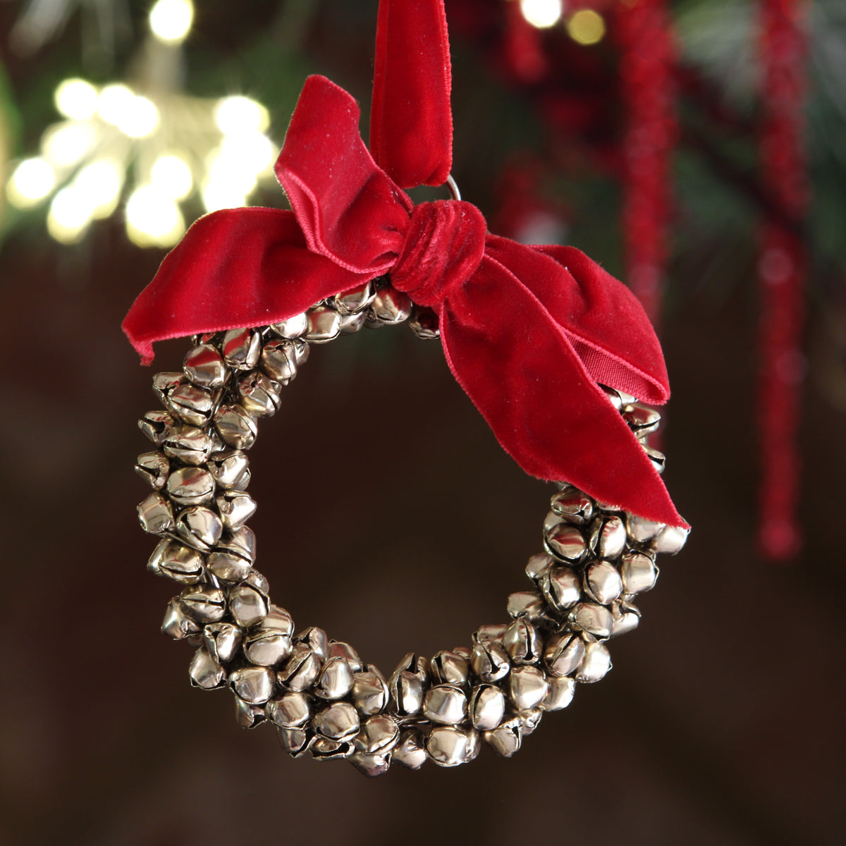 A silver jingle bell wreath Christmas ornament with a red velvet bow, hanging against a blurred festive background with warm lights.