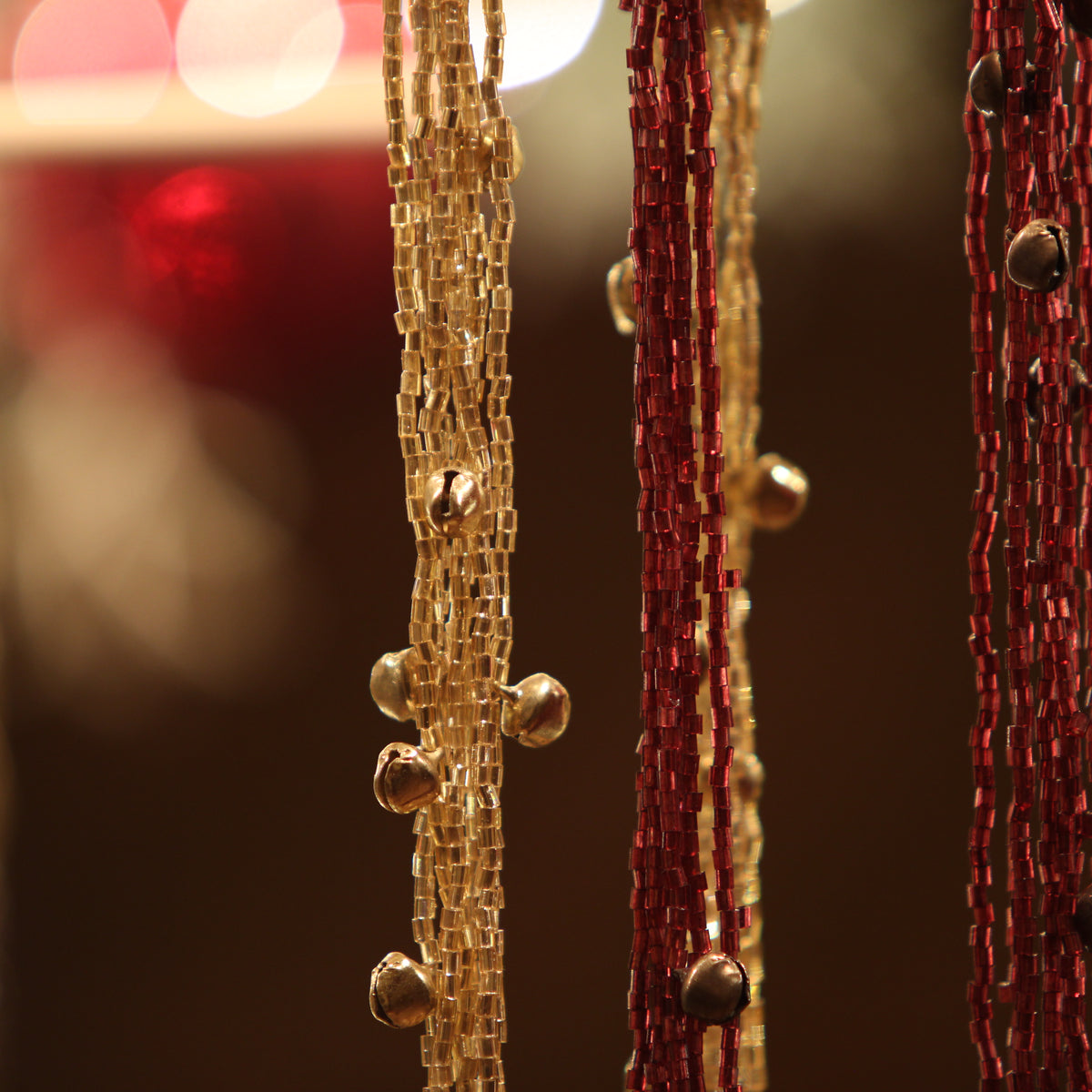 Close-up of hanging beaded strands, including a central gold-toned strand with small metallic beads and flanked by darker red and brown strands against a warm blurred background.