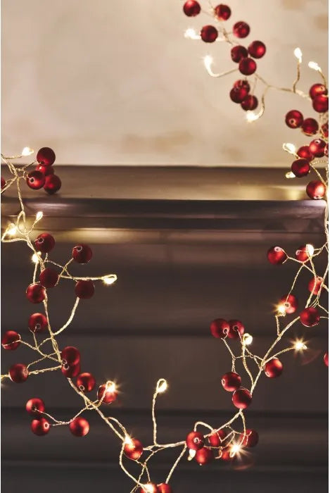 Red berry garland with warm white string lights draped around a dark mantel against a soft beige wall.