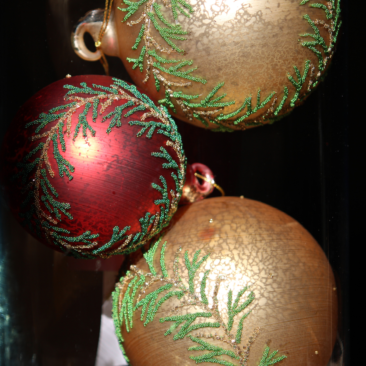 Three decorative Christmas baubles with a cracked metallic finish, featuring one red ornament and two gold ornaments, both decorated with green beaded pine branch patterns, set against a dark background.