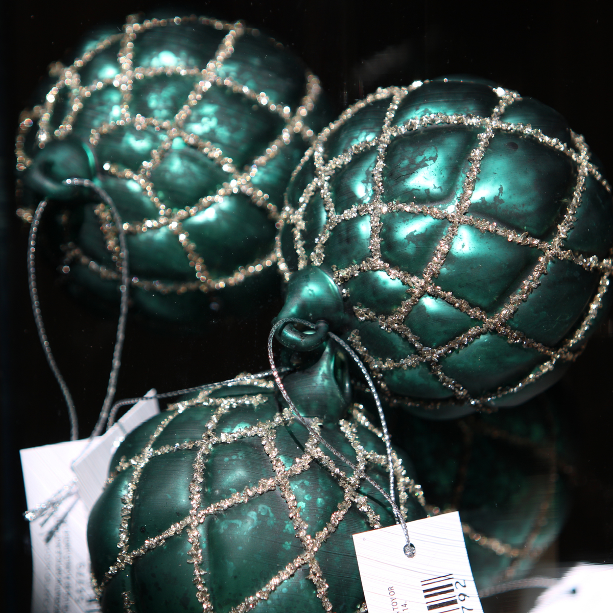 Three metallic teal green Christmas ornaments with a glittery silver lattice pattern, shown with retail tags against a dark background.