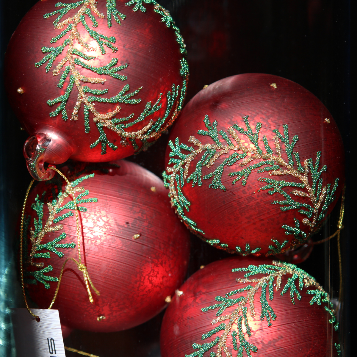 Four red Christmas ornaments with glittery gold and green pine branch patterns, shown close up against a dark background.
