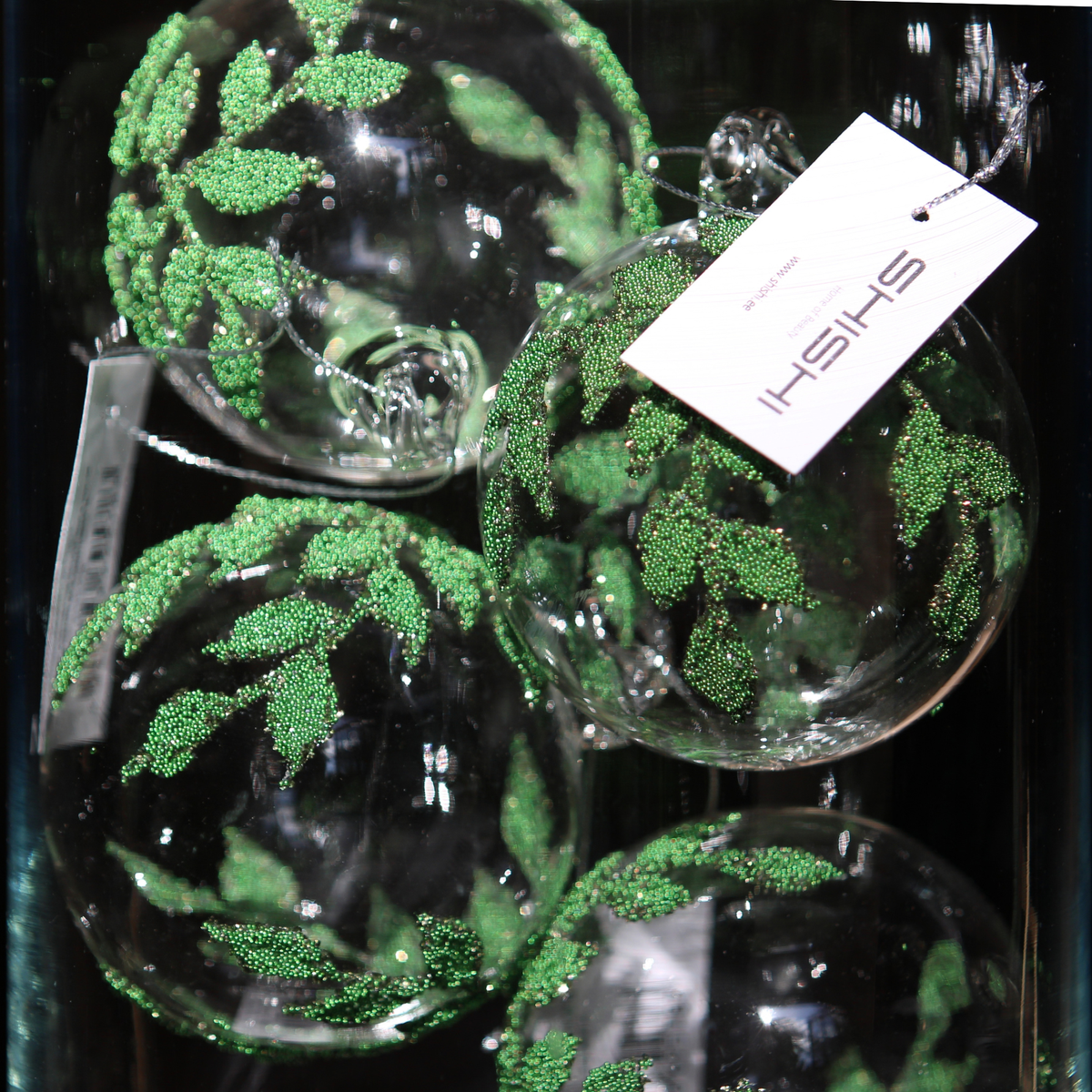 Clear glass Christmas ornaments decorated with green glittery leaf patterns, shown with a white hanging tag that reads SHISHI.