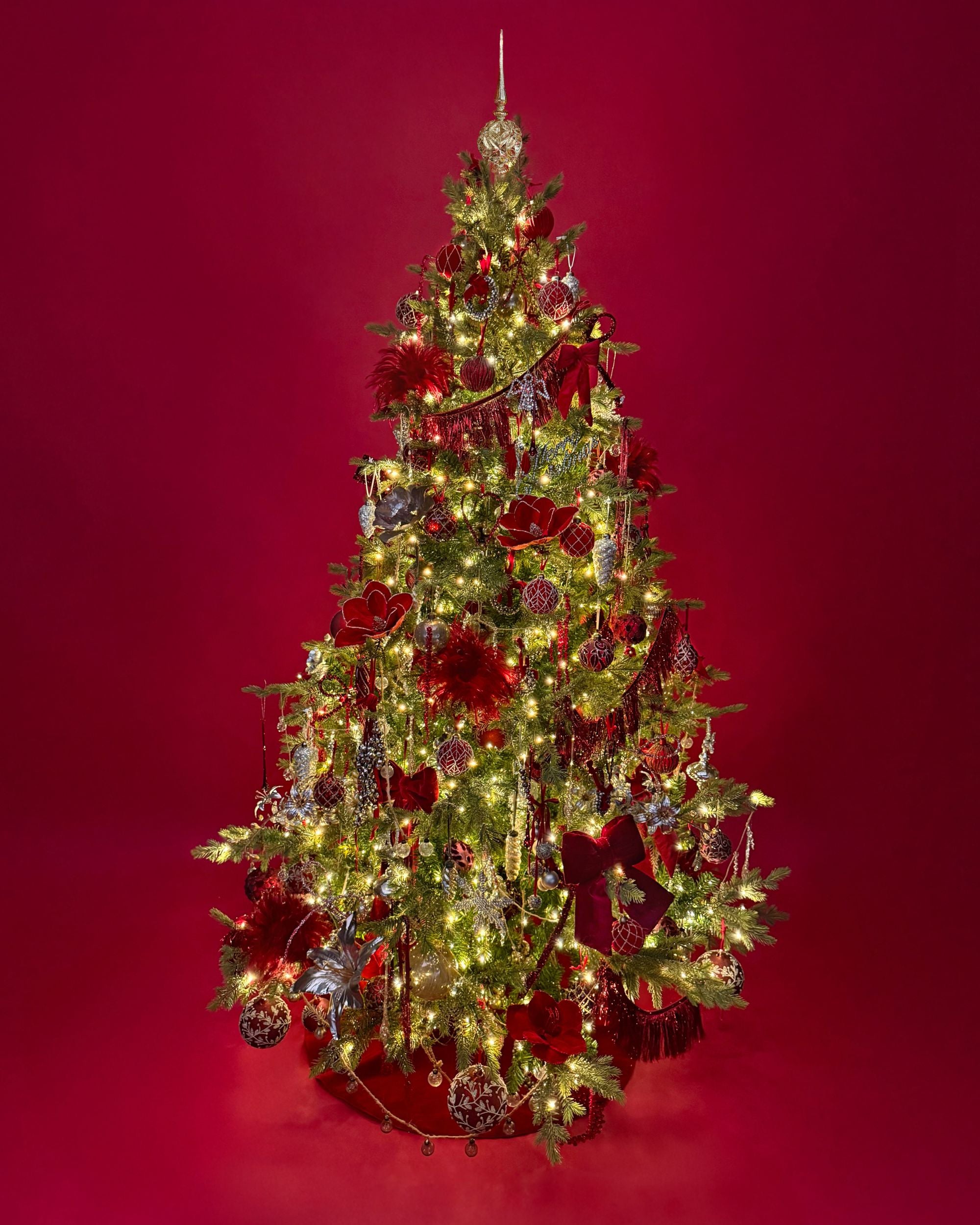 A decorated Christmas tree with green branches, warm white lights, red ribbons, and red and silver ornaments, set against a solid dark red background.