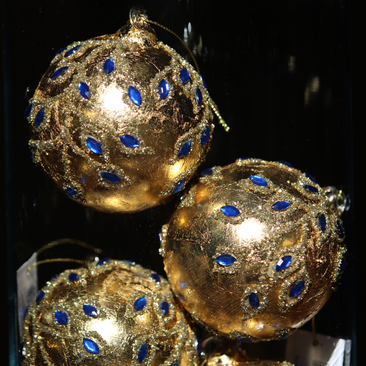 Three decorative gold Christmas tree ornaments with blue jewel accents, shown close up against a dark background.