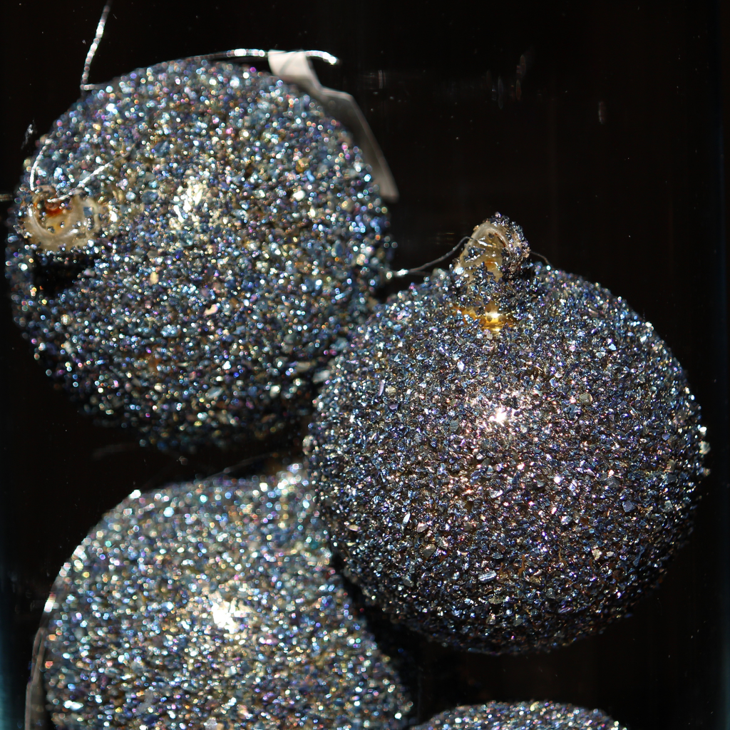 Three round glitter Christmas ornaments in a dark iridescent blue-purple color, covered in fine holographic glitter with small gold hanging loops, shown against a black background.
