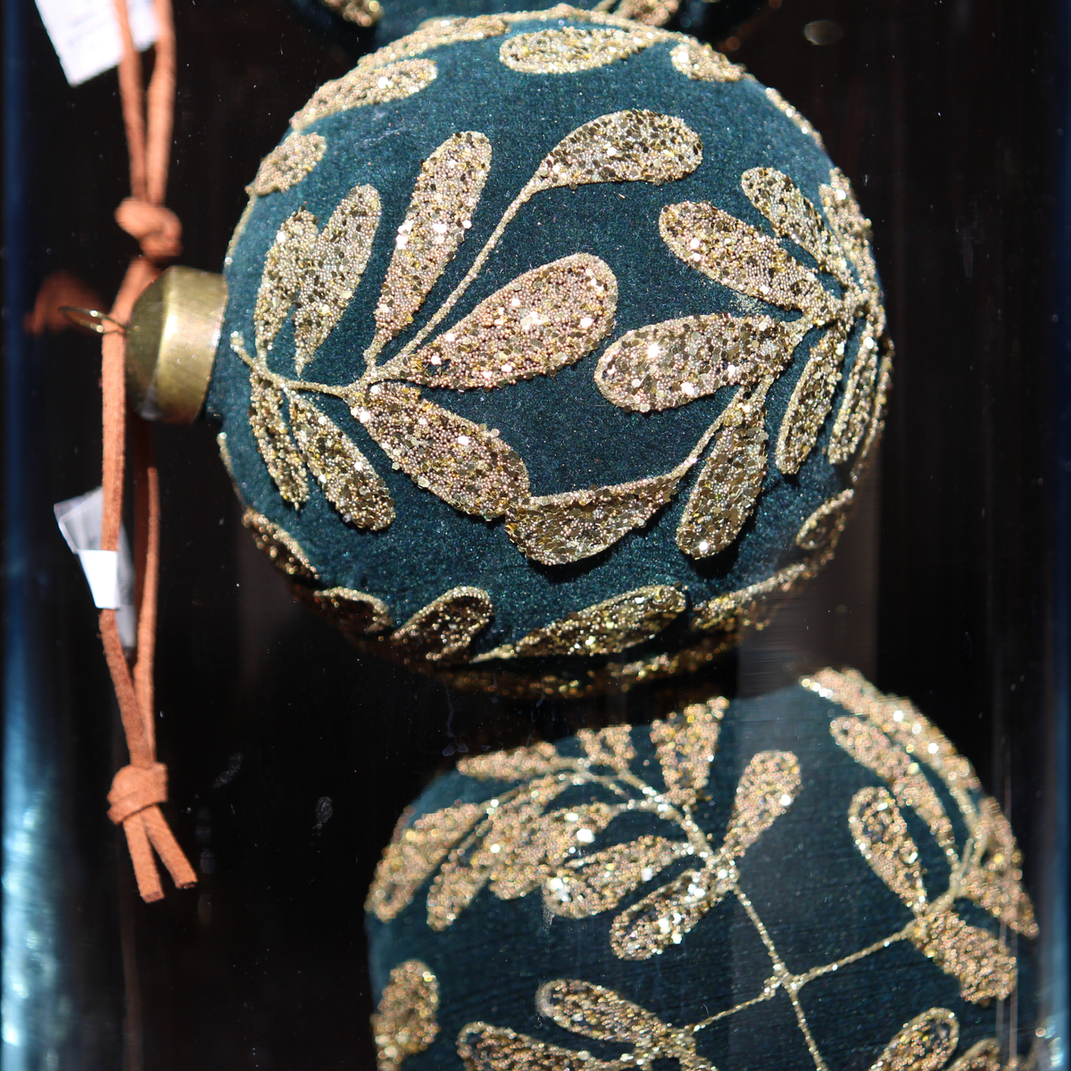 A close-up of a dark teal-green spherical ornament decorated with glittery gold leaf patterns, featuring a gold cap and an orange hanging cord.