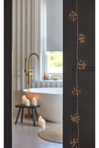 Warm white starburst fairy string lights hanging vertically on a dark wood doorframe, looking into a softly blurred modern bathroom with a white freestanding tub, gold faucet, candles, and a window.