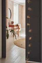 Warm white starburst string lights draped vertically down a dark charcoal door, framing a cozy interior scene with a wooden vanity, round mirror, and light-colored wood floor.