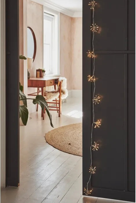 Warm white starburst string lights draped vertically down a dark charcoal door, framing a cozy interior scene with a wooden vanity, round mirror, and light-colored wood floor.