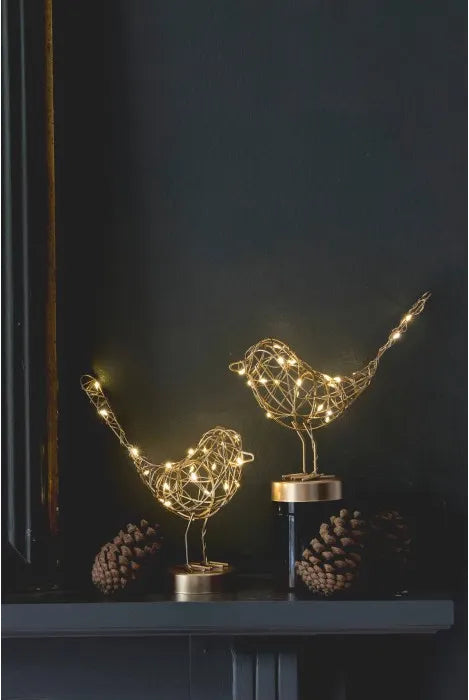 Two gold wire bird figurines lit with warm string lights, displayed on a dark mantel with pinecones against a dark wall.