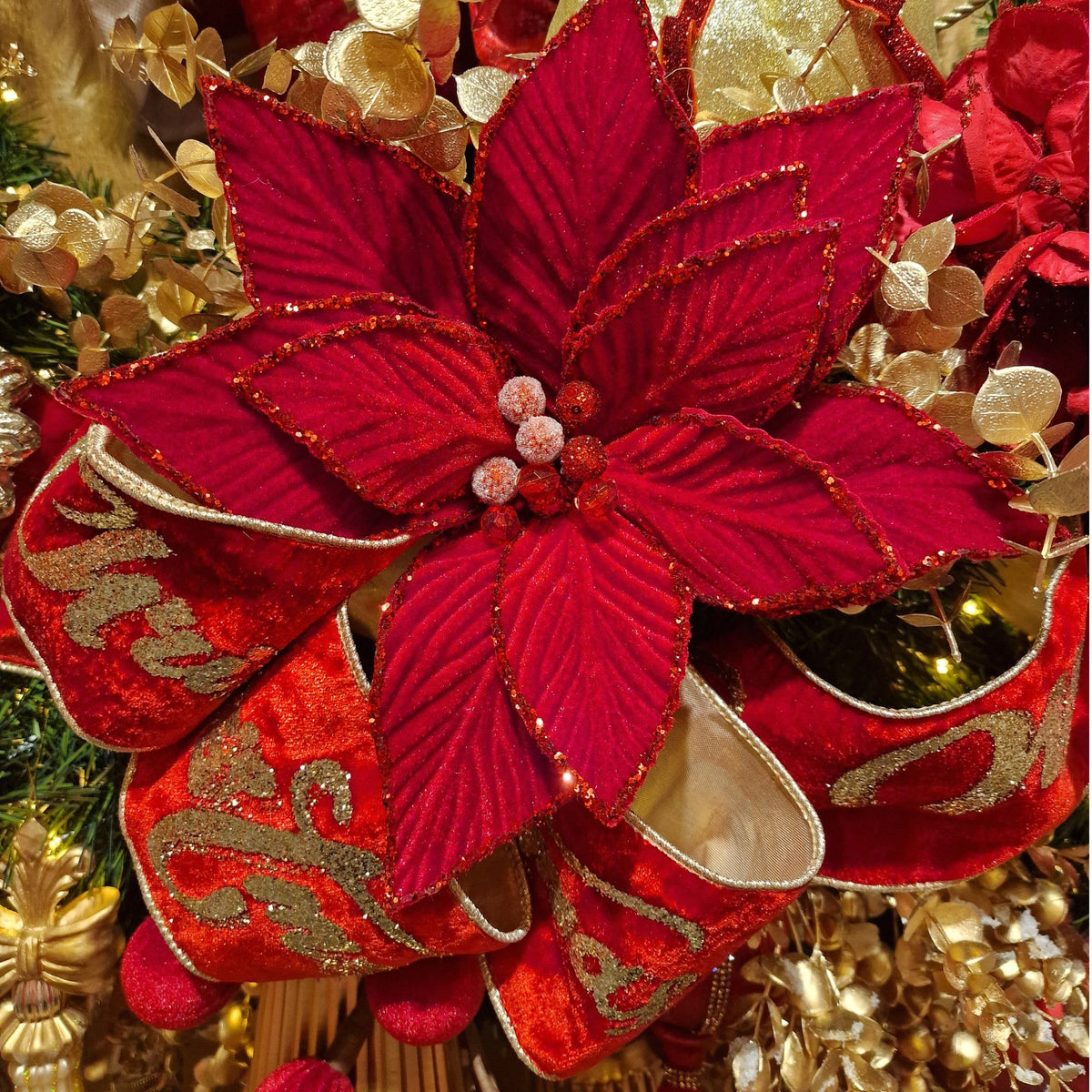 A close-up of a red poinsettia-style decorative flower ornament with glittery edges, surrounded by red and gold ribbon and gold holiday decorations.