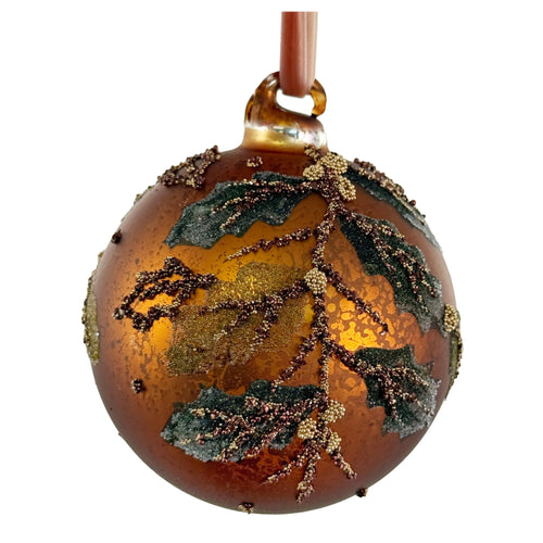 A round amber glass Christmas ornament decorated with metallic gold glitter, dark green leaf accents, and small brown bead details, hanging from a brown ribbon against a white background.