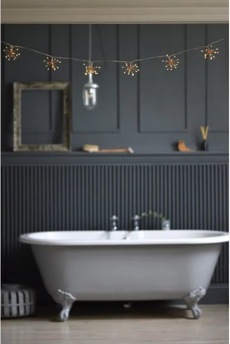 A freestanding white clawfoot bathtub in a dark charcoal bathroom with paneled walls, string lights, and a hanging glass bulb.