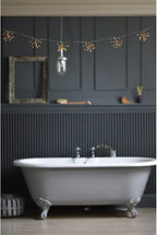 A freestanding white clawfoot bathtub in a dark charcoal bathroom with paneled walls, string lights, and a hanging glass bulb.
