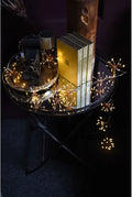 Warm white fairy string lights draped over a round black metal side table, glowing around stacked books and a vintage camera in a dark room.