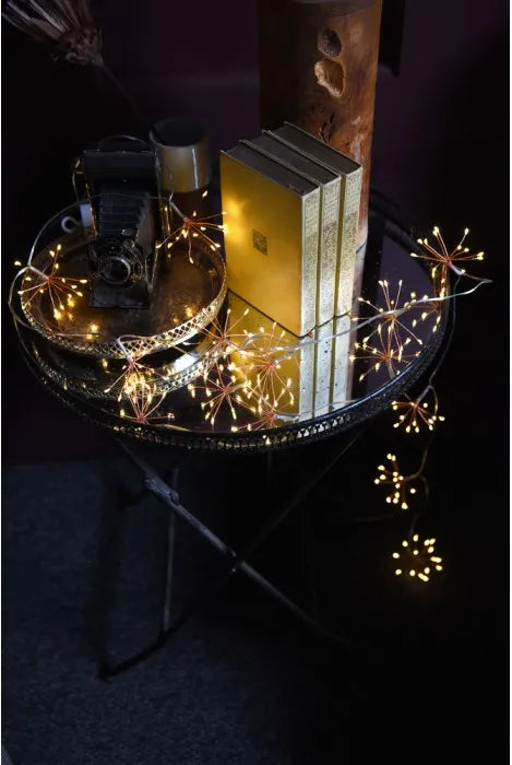 Warm white fairy string lights draped over a round black metal side table, glowing around stacked books and a vintage camera in a dark room.