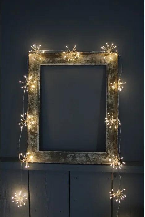 A rustic rectangular picture frame with warm white starburst string lights draped around it, set against a dark gray wall and wooden surface.